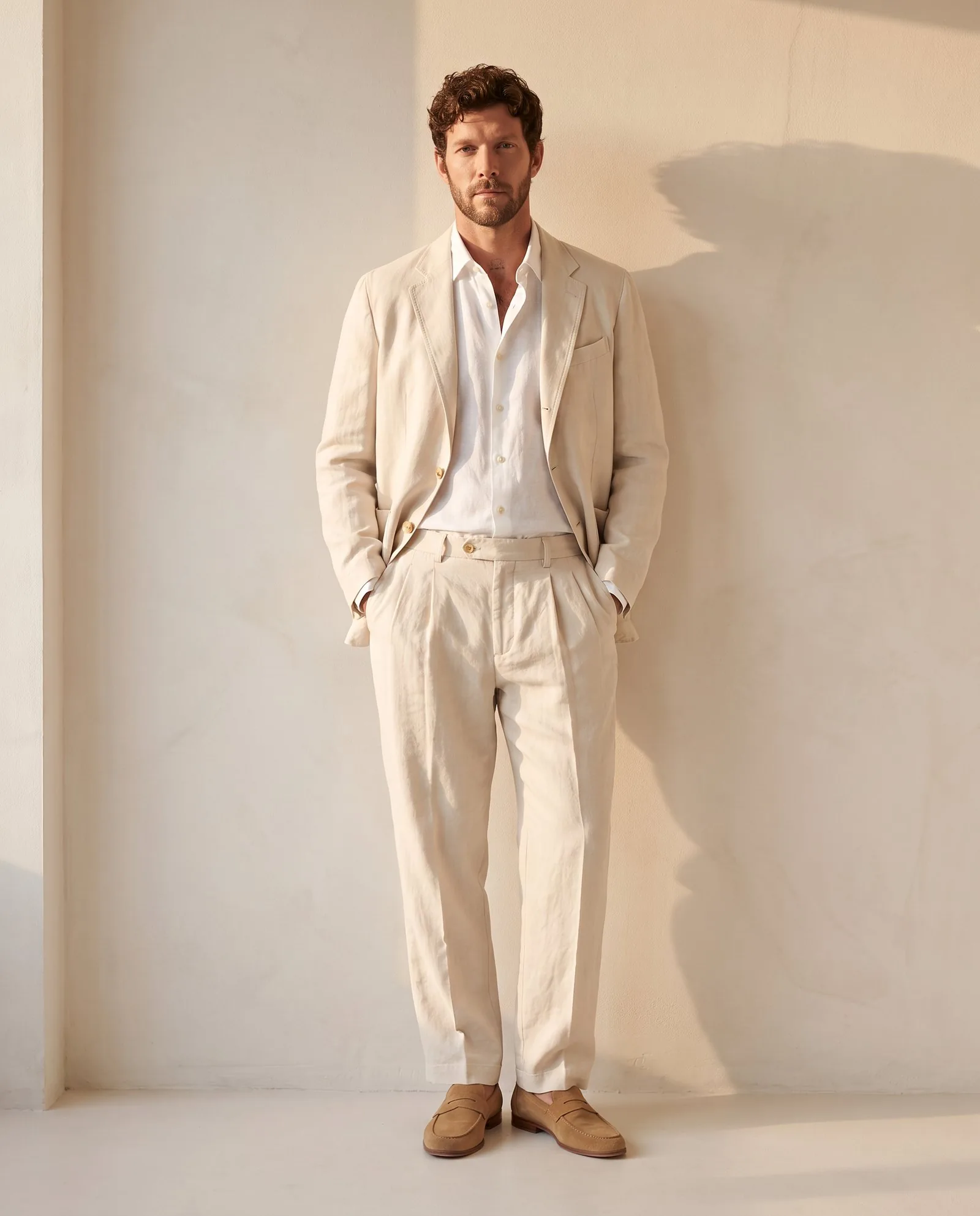 Model in a cream linen suit leaning on a sunlit plaster wall